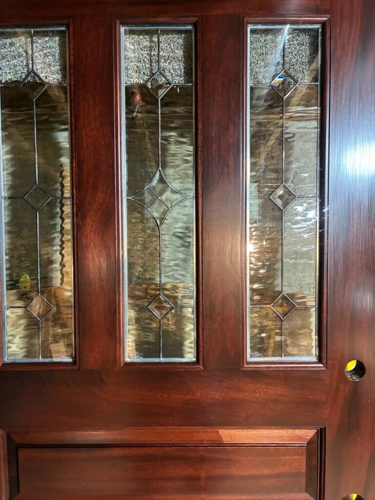 A reddish-brown wooden door featuring three vertically aligned rectangular leaded glass inserts. Each insert has a geometric pattern with a central clear diamond, surrounded by wavy clear glass, and a top section of heavily textured, pebbled glass.