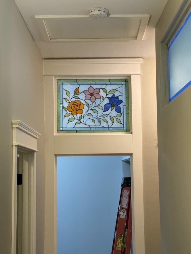 A rectangular leaded stained glass panel featuring an orange rose, a pink four-petal flower, and a blue five-petal flower with green leaves and vines on a light blue background, framed by a green geometric border.