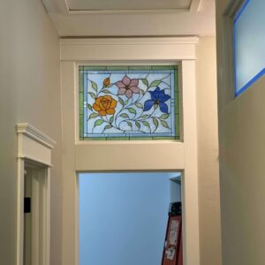 Rectangular Floral Stained Glass Panel A rectangular leaded stained glass panel featuring an orange rose, a pink four-petal flower, and a blue five-petal flower with green leaves and vines on a light blue background, framed by a green geometric border.