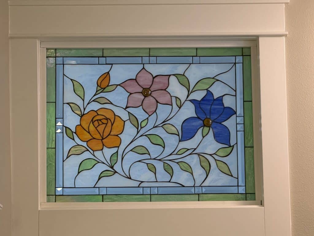 A rectangular stained glass panel featuring an orange rose, a pink four-petal flower, and a blue five-petal flower, with green leaves and vines on a light blue background, bordered by light blue beveled glass and green glass.