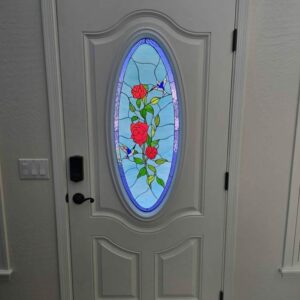 Oval Hummingbird and Rose Stained Glass Door Lite An oval, leaded stained glass door lite depicting red roses on a vine with green leaves and two blue hummingbirds against a light blue background, set into a white panel door.