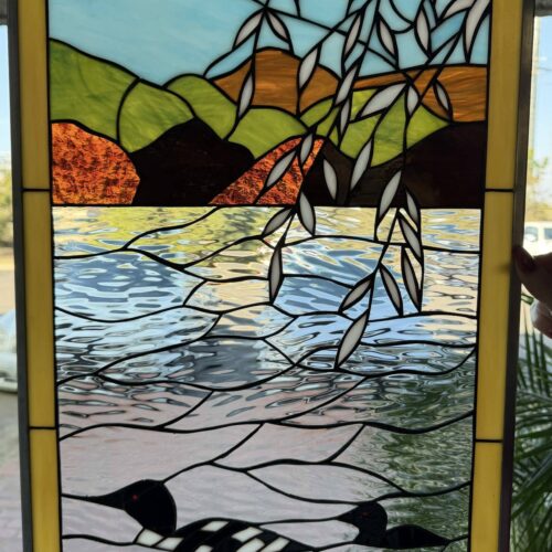 A rectangular leaded stained glass panel illustrating a serene lake landscape. Three black and white loon birds are depicted swimming in textured clear and blue water. Green and brown hills rise under a light blue sky, with white-leafed branches draped over the upper portion of the water. The panel is finished with a solid yellow border.