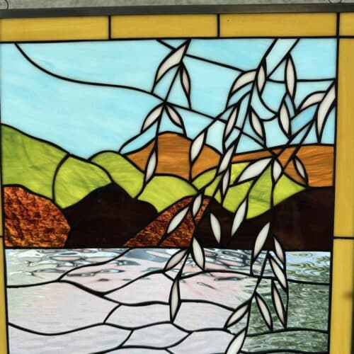 A rectangular leaded stained glass panel depicting a serene landscape. The scene features a light blue sky, rolling green and brown hills, and a shimmering water body made of textured clear and pale green glass. White leaves on hanging branches gracefully extend into the scene. The panel is bordered by yellow glass and a dark metal frame.