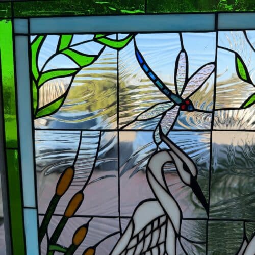 A rectangular stained glass panel depicting a vibrant wetland scene. A white egret stands gracefully among green cattails, with a blue and green dragonfly hovering above. The background features textured clear glass suggesting water and sky, bordered by light blue and green glass at the top.