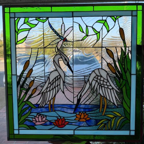 A rectangular leaded stained glass panel featuring a tranquil marshland scene. Two elegant white herons stand in blue water with pink and orange water lilies and green cattails. A blue and black dragonfly with clear wings flies above, set against a background of clear, textured glass that allows light to pass through.