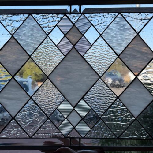 A rectangular, leaded stained glass panel with a geometric diamond pattern. It features various clear textured glass, including stippled, reamy, and linearly textured pieces, interspersed with translucent white and clear smooth diamond shapes. The panel is framed by clear beveled glass at the top, bottom, and sides.