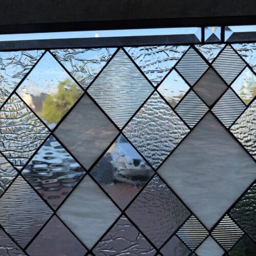 A rectangular stained glass panel with a repeating diamond grid pattern. It features various clear textured glass pieces, including hammered, reeded, and crackle, alongside translucent white opaque diamonds. The panel is framed and detailed with dark lead lines, and includes clear beveled glass accents along the top and left edges.