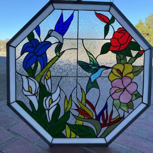 An octagonal stained glass panel depicting two hummingbirds among various colorful flowers. One blue hummingbird is near a large blue flower, while another green and light blue hummingbird is near a red rose, yellow flower, and pink flower. White calla lilies and bird of paradise flowers are also visible. The background is clear, textured glass, framed by a white glass border and a dark metal frame. Two hanging loops are visible at the top.