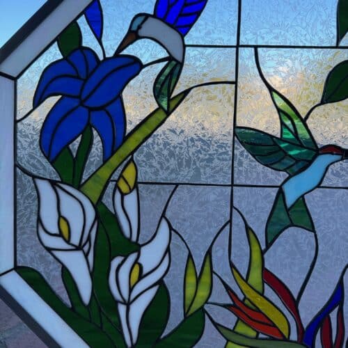A hexagonal leaded stained glass panel depicting a vibrant tropical garden scene with two hummingbirds, blue lilies, white calla lilies, and exotic red and yellow flowers, all set against a textured clear glue chip background.