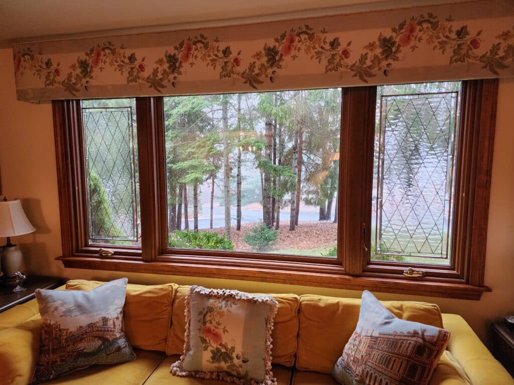 Two rectangular, leaded stained glass panels featuring a clear, textured diamond grid pattern flank a large clear window looking out onto trees and water. The panels are set within a wooden window frame.