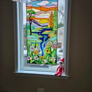 Rectangular Abstract Landscape Stained Glass Panel A rectangular, leaded stained glass panel depicting an abstract landscape. The vibrant scene includes a winding blue river, colorful wildflowers in the foreground, stylized trees, rolling hills, and a bright yellow sun against an orange and pink sky.