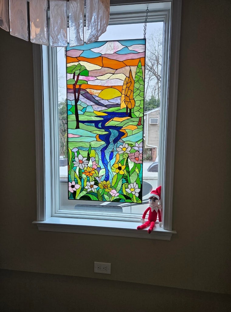 A rectangular, leaded stained glass panel depicting an abstract landscape. The vibrant scene includes a winding blue river, colorful wildflowers in the foreground, stylized trees, rolling hills, and a bright yellow sun against an orange and pink sky.