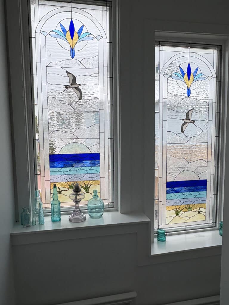 Two tall, rectangular leaded stained glass windows, each depicting a coastal scene. The top features a stylized blue and yellow floral design within clear textured glass. Below, a gray and white seagull flies over clear textured water. The lower section shows a stylized sun rising over bands of blue ocean, leading to sandy dunes with green plants.