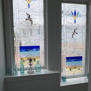 Rectangular Leaded Stained Glass Coastal Scene Panel with Seagull and Floral Motif Two tall, rectangular leaded stained glass windows, each depicting a coastal scene. The top features a stylized blue and yellow floral design within clear textured glass. Below, a gray and white seagull flies over clear textured water. The lower section shows a stylized sun rising over bands of blue ocean, leading to sandy dunes with green plants.