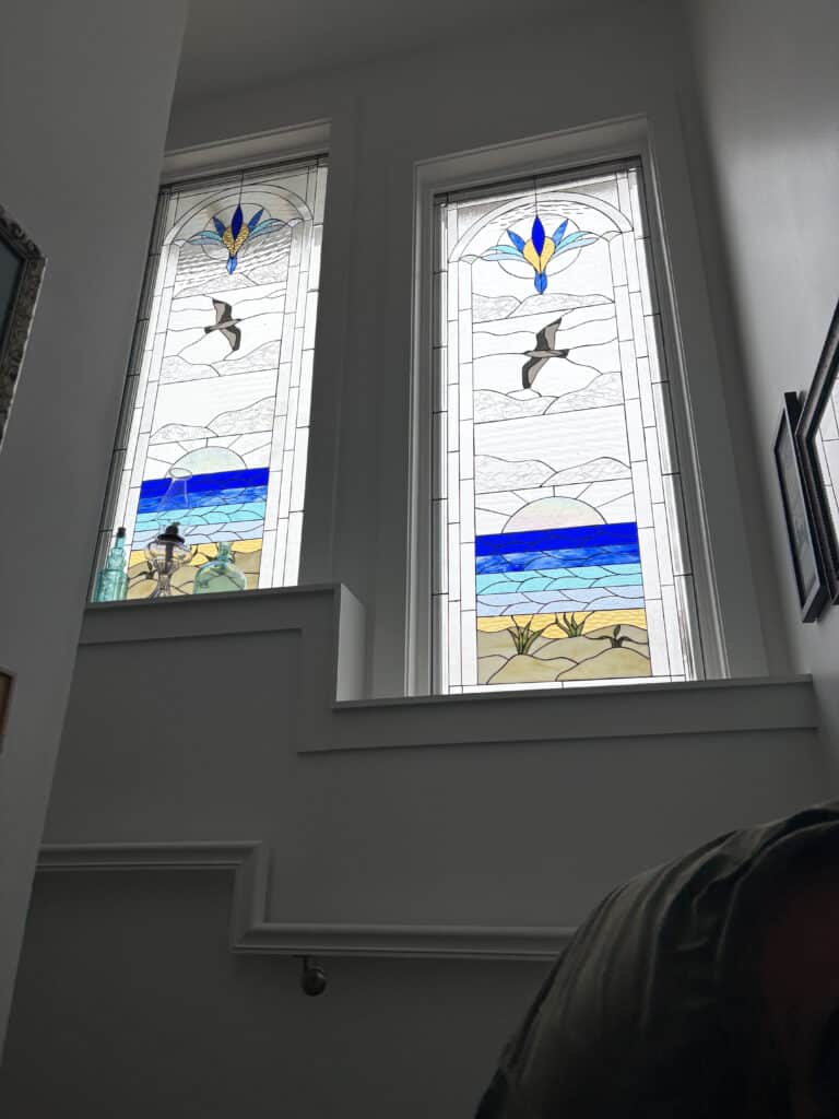 A pair of tall, rectangular leaded stained glass windows with arched tops, featuring a coastal design including sand dunes, ocean waves, a soaring bird, and a stylized floral motif in blue and yellow.