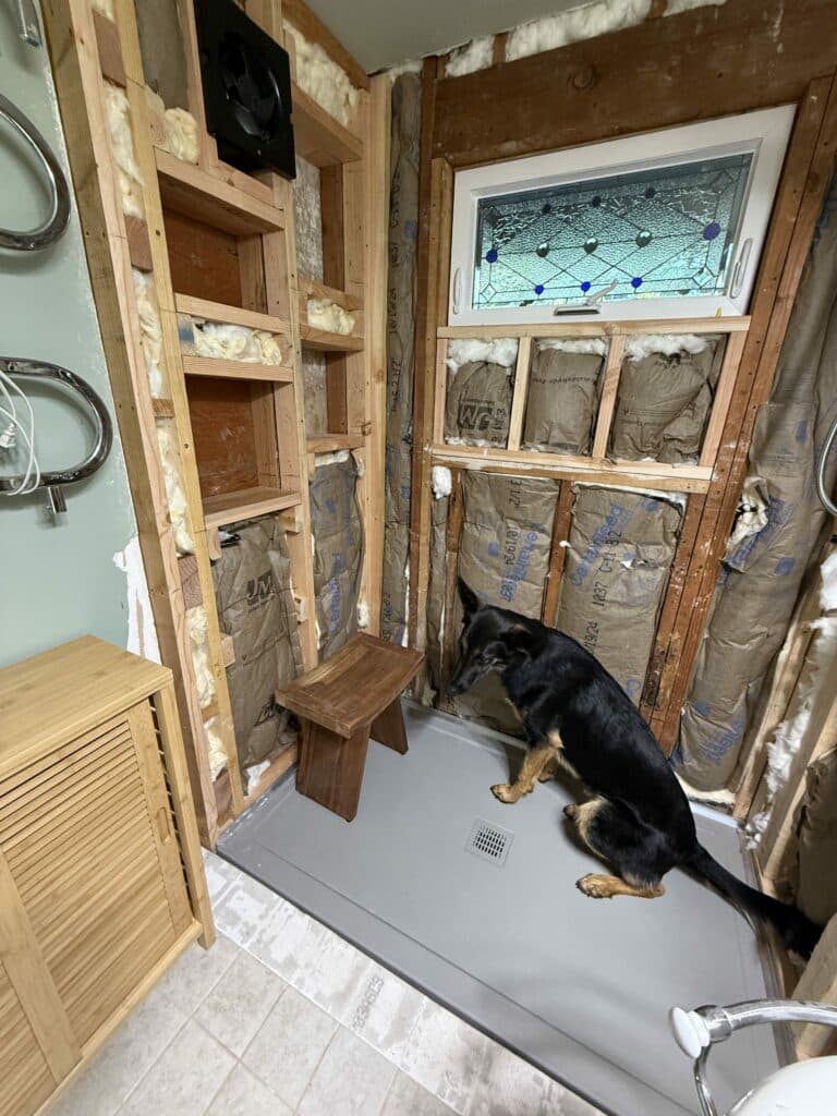 A rectangular, white-framed stained glass window with a geometric clear textured glass pattern and small blue circular accents, installed in an unfinished bathroom wall.