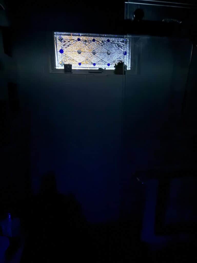 A rectangular leaded stained glass window featuring clear textured glass with a geometric pattern, accented by round and diamond-shaped blue glass jewels, set in a white frame, viewed in a dimly lit room.