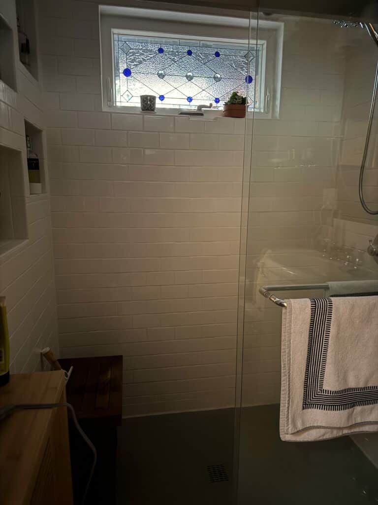 A horizontal rectangular stained glass window featuring a geometric design made of clear textured glass, clear bevels, and round cobalt blue glass jewels. The window is set in a white frame above a tiled shower.