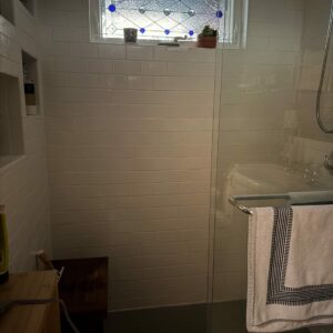 Horizontal Geometric Stained Glass Window with Blue Jewels A horizontal rectangular stained glass window featuring a geometric design made of clear textured glass, clear bevels, and round cobalt blue glass jewels. The window is set in a white frame above a tiled shower.