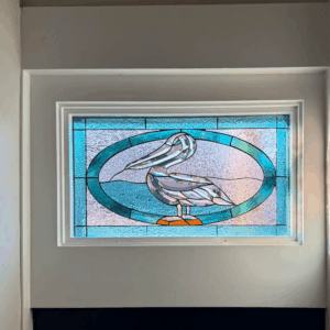 Rectangular Pelican Stained Glass Panel A rectangular, leaded stained glass panel depicting a pelican. The pelican is crafted from grey and white textured glass, standing on an orange base, centered within a teal and clear textured oval. The background consists of blue and clear textured glass.