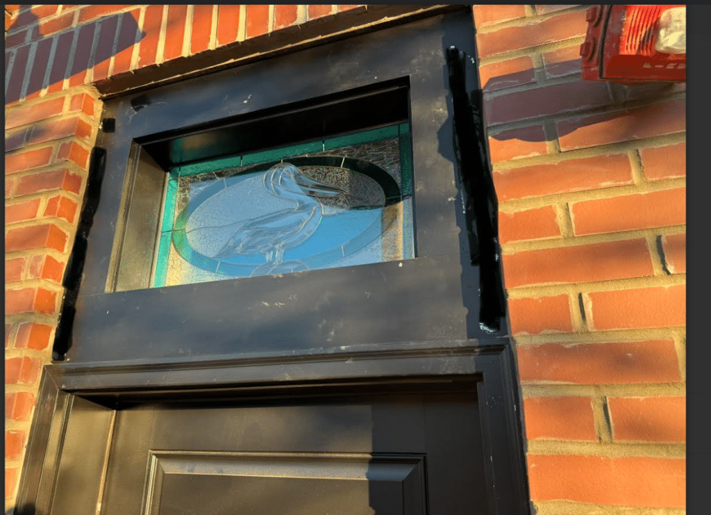 A rectangular stained glass transom panel featuring a clear etched pelican design within a central oval of blue and turquoise glass, bordered by green and surrounded by clear textured glass. The panel is set in a black frame above a black door in a brick wall.