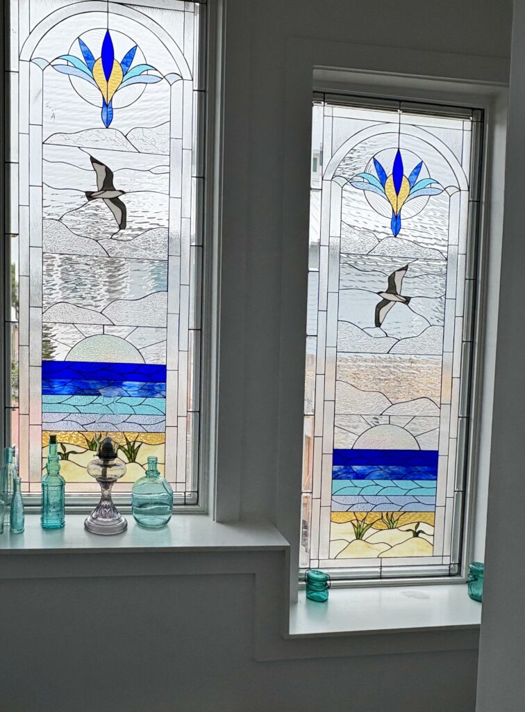 A rectangular leaded stained glass window panel depicting a coastal seascape. It features a blue and gold stylized floral design at the top, a seagull flying over textured clear glass representing the sky and ocean, a band of vibrant blue water, and sandy dunes with green plants at the bottom. This panel is shown as one of a matching pair.