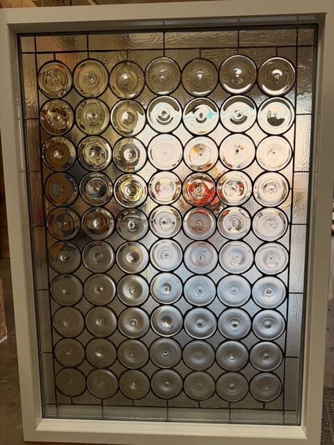 A rectangular clear stained glass window panel featuring rows of circular bullseye glass pieces surrounded by clear textured glass and set in a white frame.