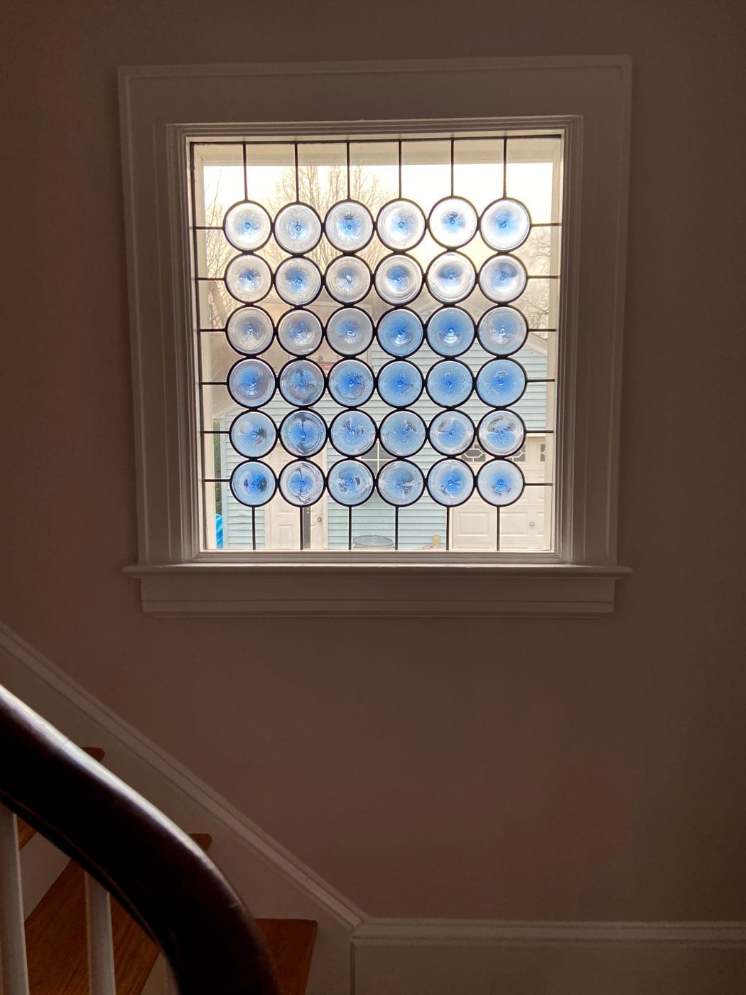 A rectangular stained glass panel featuring a grid pattern of individual clear and blue circular glass pieces, installed in a white window frame above a wooden staircase.