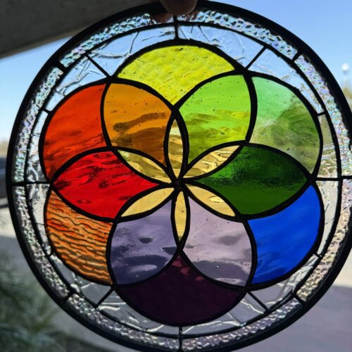 A hand holds a circular stained glass panel featuring a geometric pattern of overlapping circles in a rainbow spectrum. The central design includes vibrant red, orange, yellow, green, blue, and purple glass, while the outer ring is made of clear, iridescent textured glass. Black lead lines separate the glass pieces.