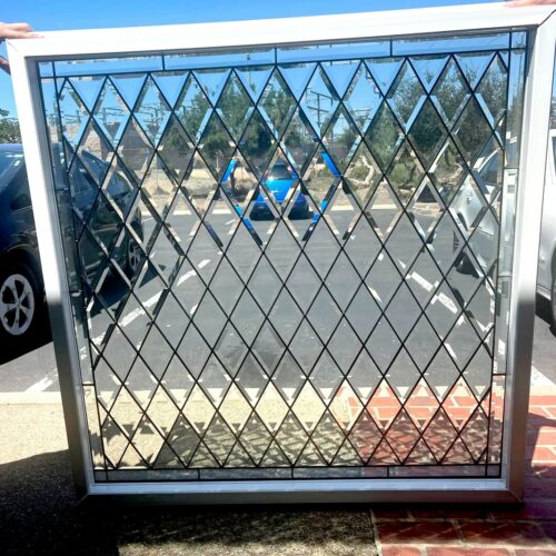 A rectangular leaded glass panel featuring an intricate pattern of clear, beveled diamond-shaped pieces. The panel is set in a white frame and is being held outdoors against a backdrop of cars and a parking lot.