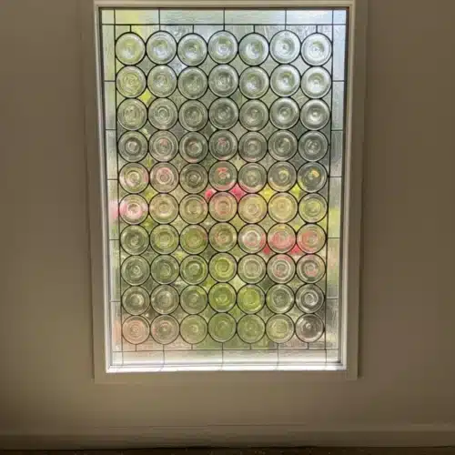 A rectangular, leaded stained glass window panel composed entirely of clear, circular bottle bottom glass pieces arranged in a grid, framed by textured clear glass.