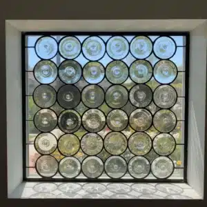 A rectangular stained glass panel made from clear, circular bullseye rondel glass pieces, set in a dark leaded grid, looking out onto a bright garden.