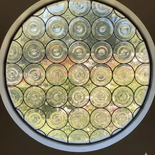 A round stained glass window made from numerous clear circular rondel glass pieces, revealing a blurred green garden scene outside.