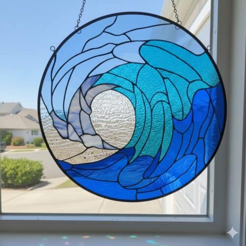 A circular stained glass panel depicting a breaking ocean wave in various shades of blue and clear textured glass, suspended in a window.