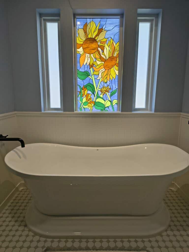 A rectangular stained glass window panel depicting vibrant yellow and orange sunflowers with green leaves against a light blue sky, installed above a white freestanding bathtub.