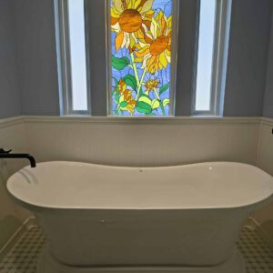 A rectangular stained glass window panel depicting vibrant yellow and orange sunflowers with green leaves against a light blue sky, installed above a white freestanding bathtub.