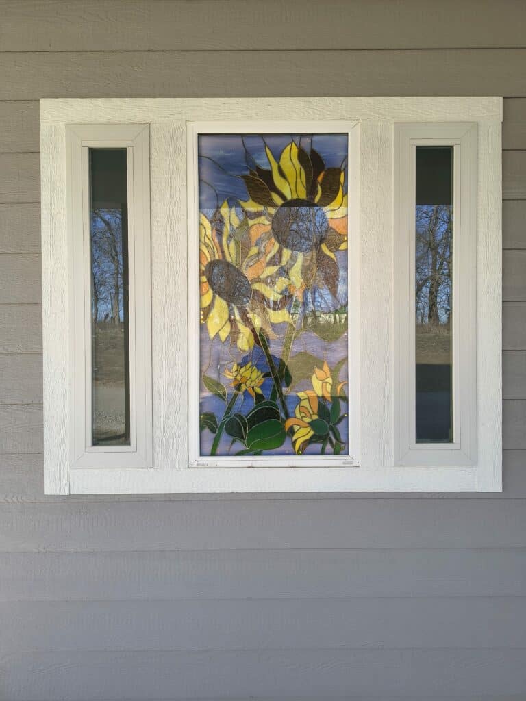 A rectangular stained glass panel depicting multiple yellow, orange, and brown sunflowers with green stems against a blue background, set within a larger window frame.