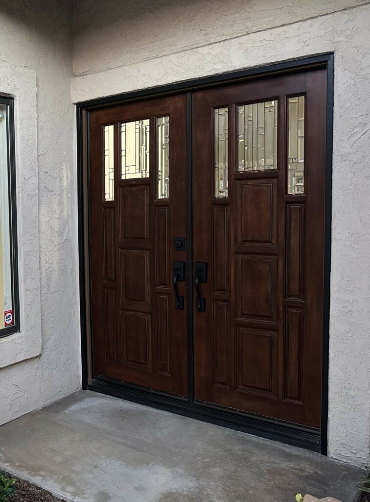 Dark brown double entry doors with black hardware, featuring clear geometric leaded glass panels in the upper sections, set in a light stucco wall.