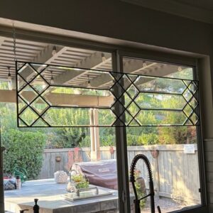 A rectangular leaded glass panel with a clear geometric pattern of rhomboid and hexagonal beveled glass pieces, suspended across the top section of a kitchen window overlooking an outdoor patio.