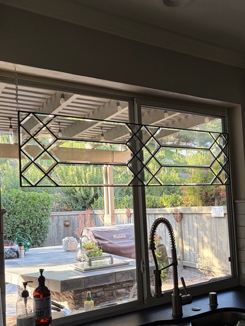 A rectangular leaded glass panel with a clear geometric pattern of rhomboid and hexagonal beveled glass pieces, suspended across the top section of a kitchen window overlooking an outdoor patio.