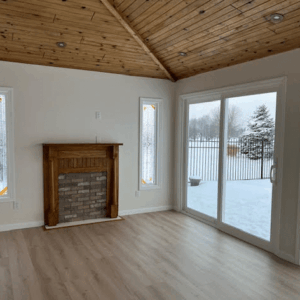 Two tall, narrow rectangular stained glass panels with a clear textured and beveled glass geometric design, flanking a wooden fireplace. The room features a vaulted wood ceiling and light wood floors, with a snowy winter scene visible outside.