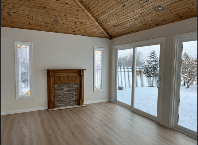 Two tall, narrow rectangular stained glass panels with a clear textured and beveled glass geometric design, flanking a wooden fireplace. The room features a vaulted wood ceiling and light wood floors, with a snowy winter scene visible outside.
