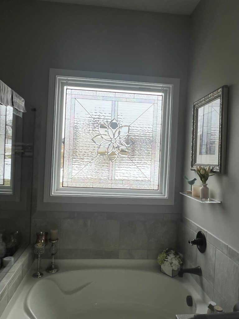 A square, leaded stained glass window with a central clear beveled floral medallion design, surrounded by clear textured glass. The window is installed in a white frame above a bathtub.