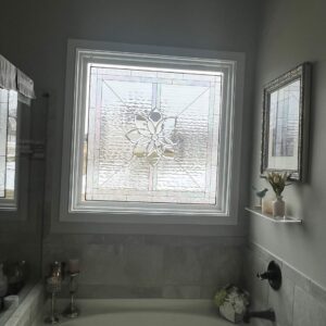 A square, leaded stained glass window with a central clear beveled floral medallion design, surrounded by clear textured glass. The window is installed in a white frame above a bathtub.
