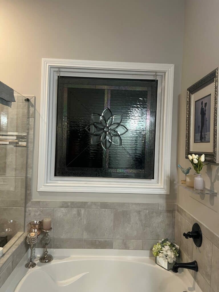 A rectangular leaded stained glass panel featuring a prominent central clear beveled flower medallion, surrounded by textured clear glass and a darker iridescent border, installed in a white window frame above a white bathtub in a modern bathroom.