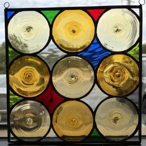 A square, leaded stained glass panel with a 3x3 grid of nine clear and golden circular rondels. The panel features red, blue, green, and clear geometric filler glass and is seen hanging in a window.