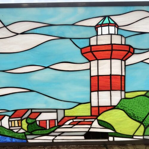 A rectangular stained glass panel depicting a coastal scene with a red and white striped lighthouse, houses, green land, blue water, and a blue sky with white clouds.