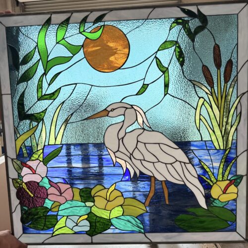 A rectangular leaded stained glass panel in a metal frame, featuring a white egret standing in blue water. The scene includes green marsh plants, pink, yellow, and purple flowers, brown cattails, and an orange sun against a textured blue sky.
