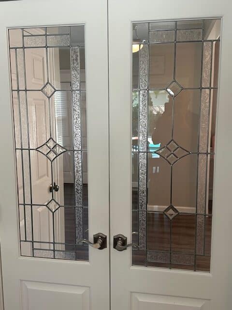 A pair of white double doors, each with a rectangular clear leaded glass insert. The inserts feature a geometric pattern of clear beveled glass diamonds and textured vertical panes.